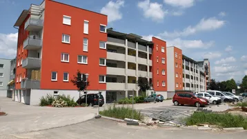 Modernes Mehrfamilienhaus mit leuchtend roter Fassade, Balkonen und umliegenden Parkplätzen unter blauem Himmel.