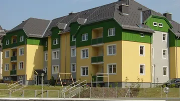 Mehrfamilienhaus mit gelb-grüner Fassade, Balkonen, Spielplatz und Garagen im Vordergrund.