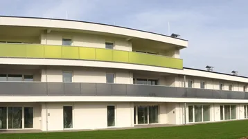 Modernes Mehrfamilienhaus mit weißer Fassade, großen Fenstern und farbigen Balkonen, umgeben von einer Rasenfläche.