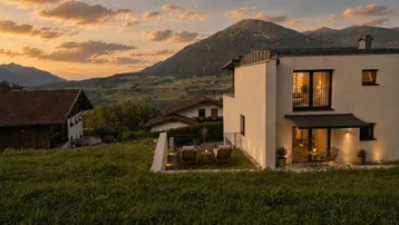 Modernes Haus mit beleuchteter Terrasse und Liegestühlen, umgeben von grünen Bergen bei Sonnenuntergang.