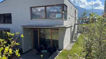 Rückseite des Hauses mit Holzterrasse, Garten und Blick auf die Berge in der Ferne.