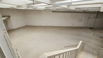 Großer, leerer Raum mit Betonboden und sichtbaren Deckenbalken, der über eine Treppe zugänglich ist.