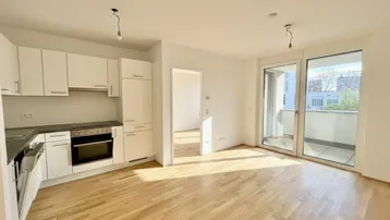Heller offener Wohnbereich mit integrierter Küche, Parkettboden und Zugang zum Balkon.