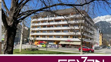 Mehrstöckiges Wohngebäude mit gelber Fassade, Balkonen und Geschäften im Erdgeschoss, umgeben von Bäumen und Bergen im Hintergrund.