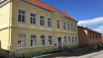 Gelbes Mehrfamilienhaus mit rotem Ziegeldach und weißen Fenstern an einer Straße gelegen.