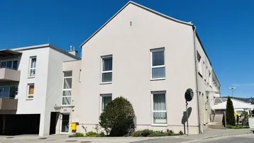 Helle Fassade eines modernen Wohngebäudes mit Satteldach und gepflegter Umgebung unter blauem Himmel.