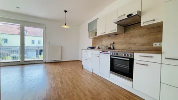 Heller offener Wohn-Essbereich mit integrierter Küche, Laminatboden und Zugang zum Balkon.