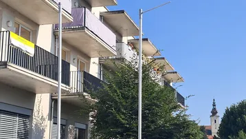 Nahaufnahme der Balkone eines modernen Wohnhauses mit grünen Hecken und einem Kirchturm in der Ferne.