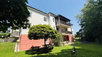 Gebäudeansicht mit Gartenbereich, Picknicktisch und Spielgeräten unter einem Baum.