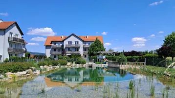 Natürlicher Schwimmteich mit klarem Wasser und Wohngebäuden im Hintergrund unter blauem Himmel.