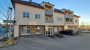 Mehrfamilienhaus mit Geschäftsräumen im Erdgeschoss und Parkplätzen davor unter klarem Himmel.