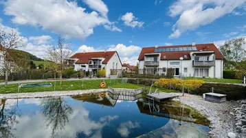 Moderne Wohngebäude mit Solaranlagen, umgeben von einer gepflegten Grünfläche und einem Naturteich unter blauem Himmel.