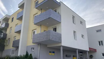 Modernes Mehrfamilienhaus mit gelber und weißer Fassade sowie grauen Balkonen unter bewölktem Himmel.