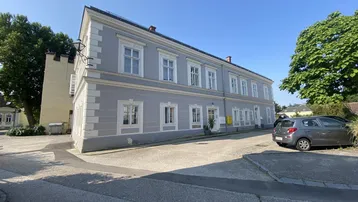 Historisches Gebäude mit grauer Fassade und weißen Fensterrahmen an einem sonnigen Tag.