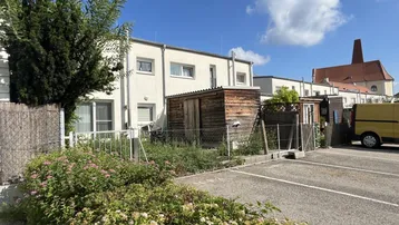Rückseite eines Wohngebäudes mit kleinem Gartenbereich, Holzschuppen und angrenzendem Parkplatz unter blauem Himmel.