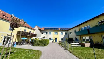 Mehrere Gebäude umgeben einen gepflegten Innenhof mit Grünflächen und einem Weg unter blauem Himmel.