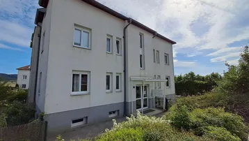 Weißes Mehrfamilienhaus mit grauer Sockelzone und Fenstern unter blauem Himmel und grüner Umgebung.