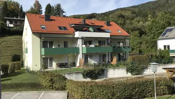 Mehrfamilienhaus mit rotem Ziegeldach, Balkonen und umgebender Natur in sonniger Hanglage.