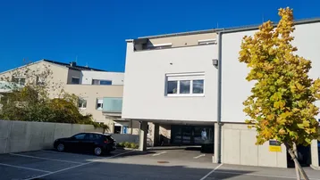 Moderne Gebäudefassade mit Außenparkplätzen und Herbstbaum unter blauem Himmel.