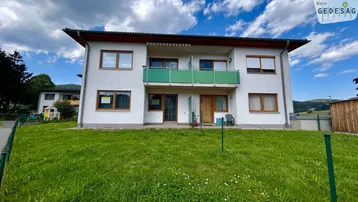Mehrfamilienhaus mit weißer Fassade, Balkon und grünem Vorgarten unter blauem Himmel.