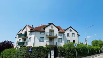 Mehrfamilienhaus mit weißer Fassade, grünen Balkonen und gepflegter Hecke unter blauem Himmel.
