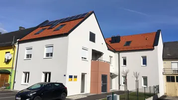 Modernes Mehrfamilienhaus mit weißer Fassade, rotem Ziegeldach und Photovoltaikanlage.