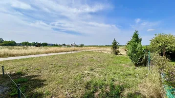 Weitläufige Landschaft mit Feldern, einem Weg und vereinzelten Bäumen unter blauem Himmel.