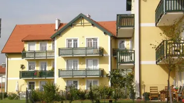 Gelbes Mehrfamilienhaus mit roten Ziegeldächern und grünen Balkonen, umgeben von einem gepflegten Garten.