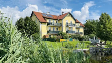 Gelbes Mehrfamilienhaus mit roten Ziegeldächern und Balkonen, umgeben von üppigem Grün und einem Teich.