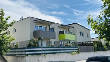 Modernes Mehrfamilienhaus mit hellen Fassaden, farbigen Balkonen und grüner Umgebung unter blauem Himmel.