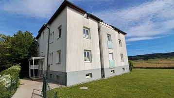 Weißes Mehrfamilienhaus mit grüner Wiese und Blick auf die umliegende Landschaft unter blauem Himmel.