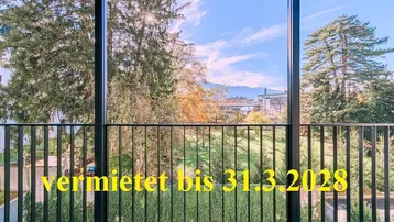 Panoramablick von einem Balkon mit Blick auf grüne Bäume, städtische Gebäude und ferne Berge unter blauem Himmel.