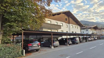 Mehrfamilienhaus mit Carport-Parkplätzen und herbstlicher Vegetation vor einer Bergkulisse.