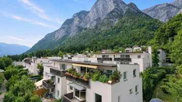 Moderne Wohngebäude mit Balkonen und Flachdächern, eingebettet in eine grüne Landschaft mit beeindruckendem Bergblick.