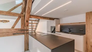 Moderne Küche mit Kochinsel, integrierten Geräten und freiliegenden Holzbalken in einem offenen Raum.