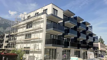 Modernes Mehrfamilienhaus im Bau mit Gerüst und Balkonen, umgeben von Bergen.
