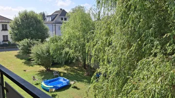 Blick vom Balkon auf den Gemeinschaftsgarten mit Bäumen und einem kleinen Planschbecken unter blauem Himmel.