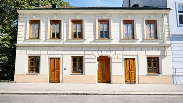 Gut erhaltene Altbaufassade mit traditionellen Holzfenstern und Holztüren, umgeben von Grün.