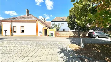 Ansicht des Gebäudes von der Straße mit gepflastertem Vorplatz und umliegenden Häusern unter blauem Himmel.