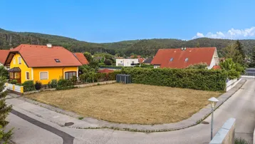 Luftaufnahme eines unbebauten Grundstücks an einer Straßenecke, umgeben von Wohnhäusern und grünen Hügeln.