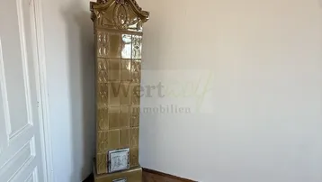 Helles Zimmer mit Fischgrätparkettboden und einem dekorativen Kachelofen in der Ecke.
