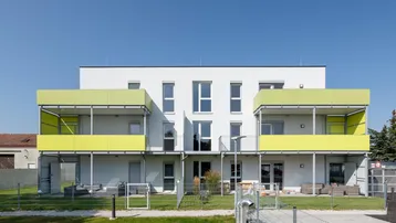 Modernes Mehrfamilienhaus mit leuchtend grünen Balkonen und Terrassen im Erdgeschoss, umgeben von Grünflächen.