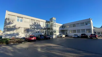 Modernes Bürogebäude mit mehreren Etagen, großen Fenstern und einem gläsernen Aufzug im Eingangsbereich.