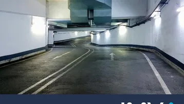 Steile Zufahrtsrampe in einer Tiefgarage mit hellen Wandleuchten und sichtbaren Lüftungskanälen an der Decke.