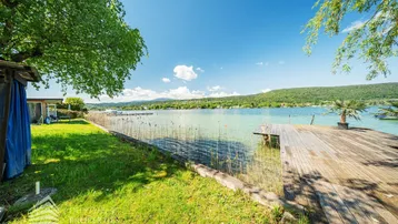 Seeufer mit Holzsteg, grüner Wiese und klarem Wasser unter blauem Himmel.