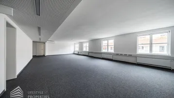 Geräumiges, leeres Büro mit grauem Teppichboden, weißen Wänden und mehreren Fenstern für natürliches Licht.