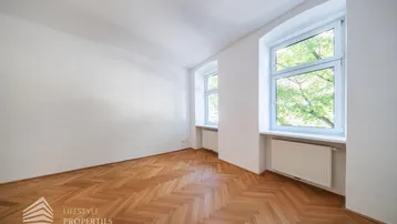 Helles Zimmer mit Fischgrätparkettboden und zwei Fenstern, die viel Tageslicht hereinlassen.