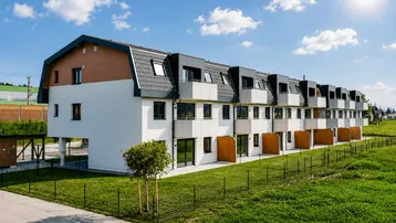 Moderne Reihenhäuser mit Satteldach und Balkonen, umgeben von grünen Wiesen unter blauem Himmel.