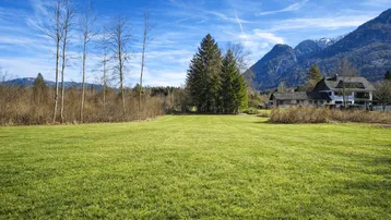 Weitläufige Grünfläche mit Blick auf ein Wohnhaus und die umliegende Berglandschaft unter blauem Himmel.