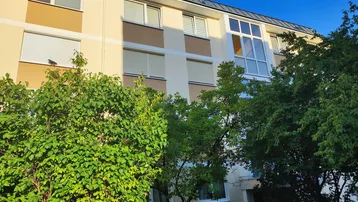 Mehrfamilienhaus mit mehreren Etagen, Fenstern mit Rollläden und begrüntem Vorgarten.